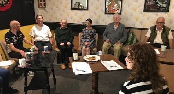 Seven people sat around a table in an informal setting with paper and biscuits on the table