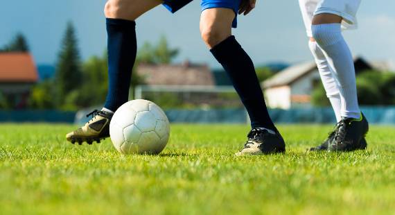 Two people playing football on grass. Shows the lower part of their legs with a football in the foreground.